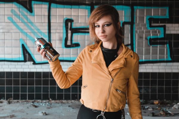 Imagem de uma mulher com um spray de tinta na mão e uma parede pichada no fundo simbolizando o arquétipo fora da lei.